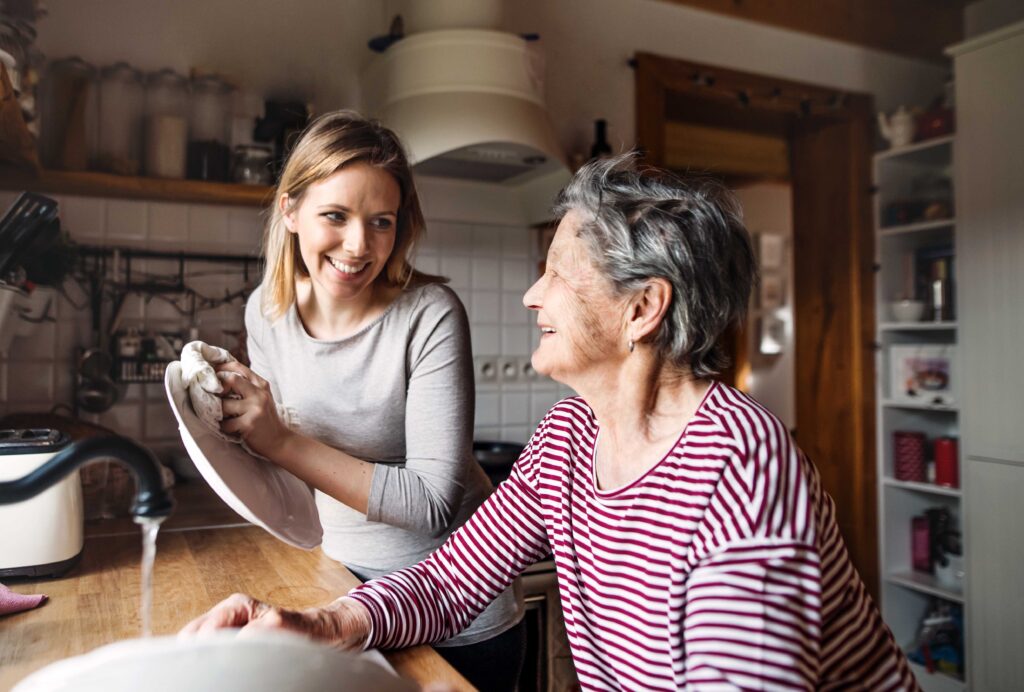 Young woman washes dishes with elderly parent in the kitchen; smiling - Alles Law